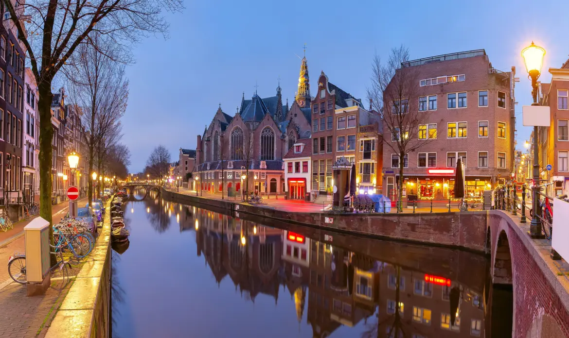 The Oude Kerk rising above a canal in Amsterdam's Red Light District at dusk, with red-lit windows reflected in the water