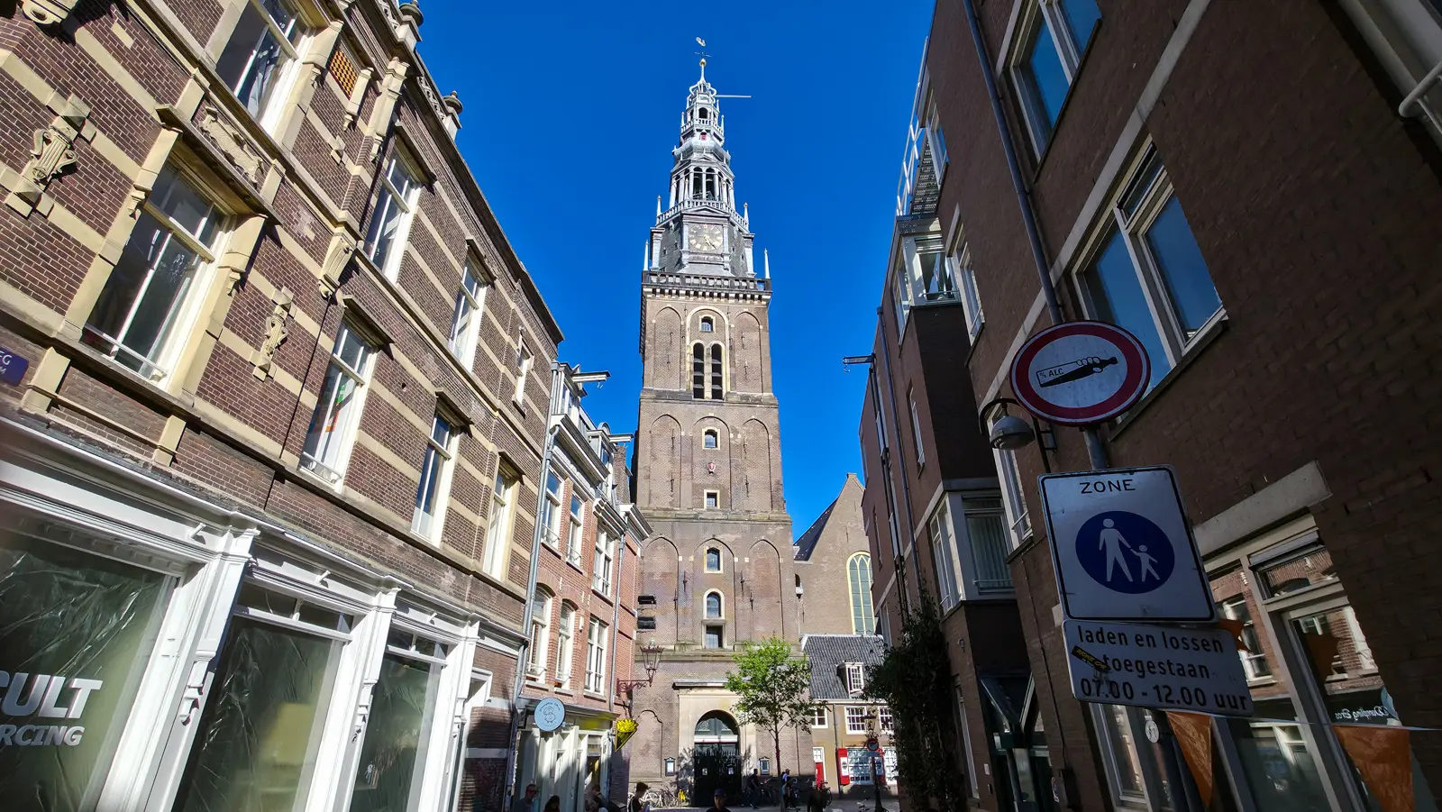 The tower of the Oude Kerk in Amsterdam against a blue sky, with a no-alcohol sign and a pedestrian zone sign in front