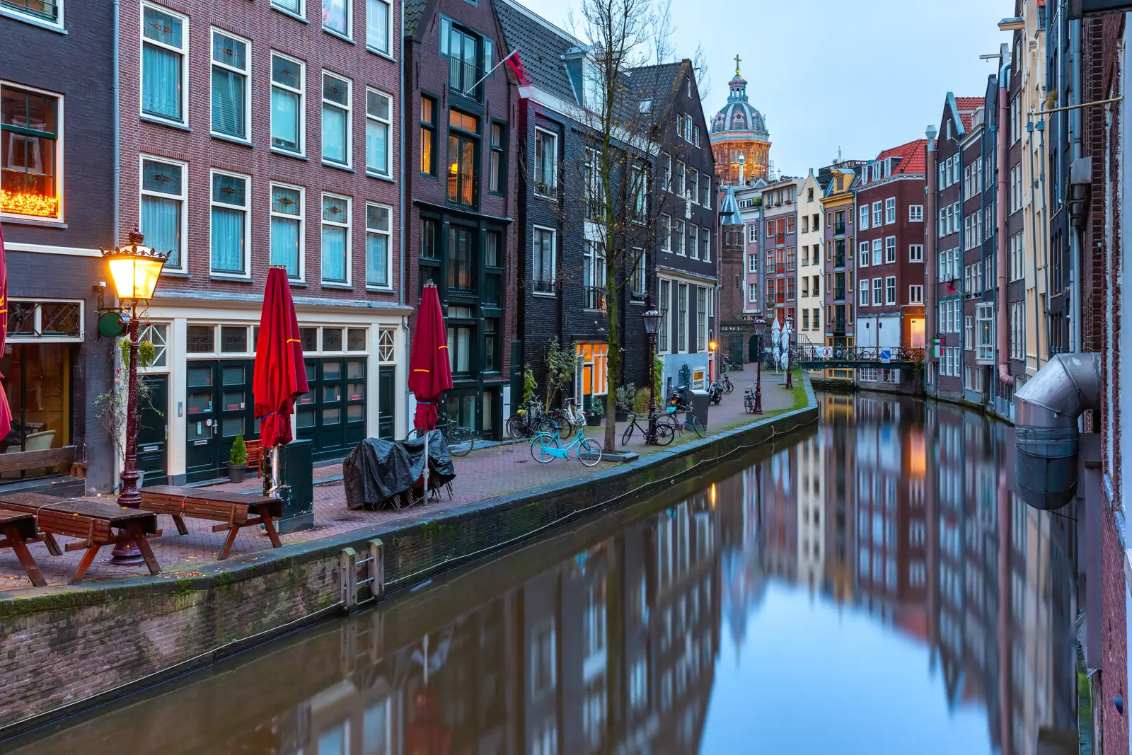 A quiet canal near the Red Light District at dawn, with red umbrellas, parked bikes and a church dome in the distance