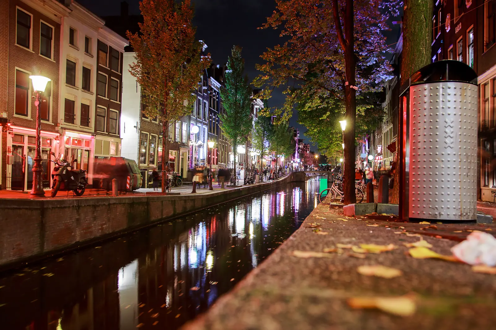 A canal in the Red Light District at night, with red and purple lights reflected in the water and people walking the quays