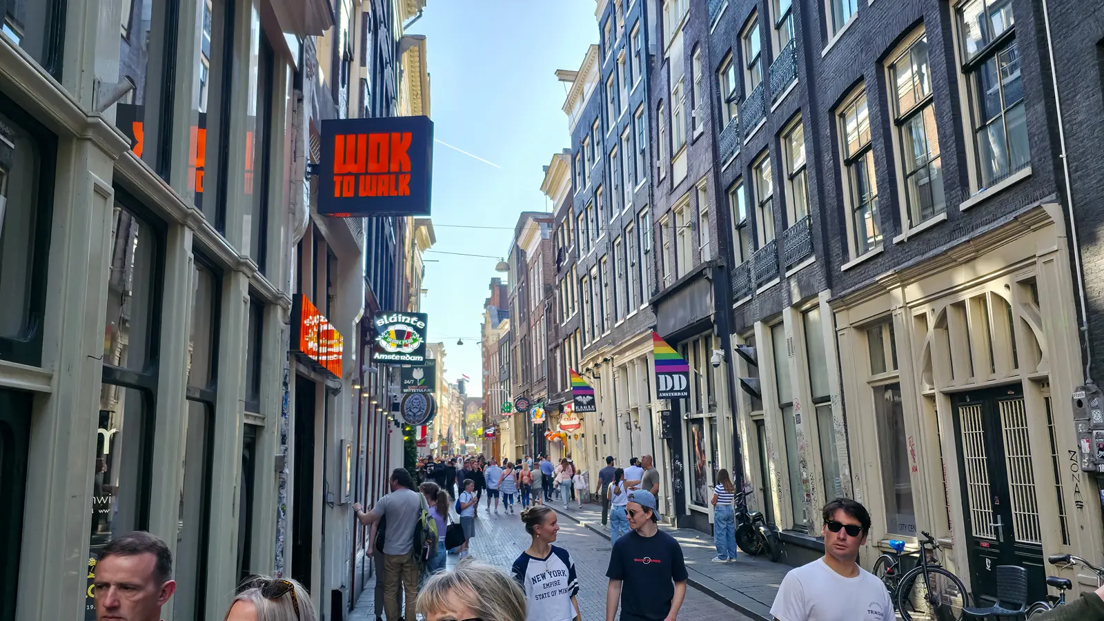 A busy shopping street on the edge of the Red Light District in Amsterdam, with pubs, food signs and rainbow flags