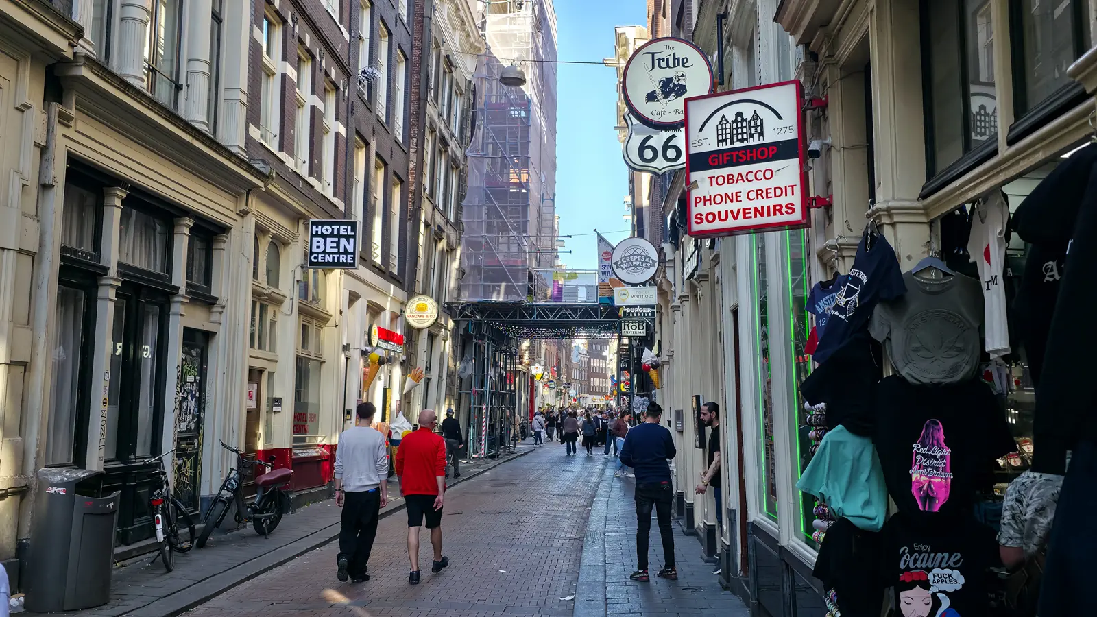 A narrow street near the Red Light District with souvenir T-shirts, tobacco and gift shop signs, and a hotel sign