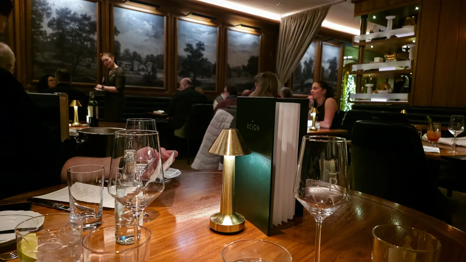 Table with a brass lamp, a green Reign menu and wine glasses, with diners and a server in the dining room behind, Toronto