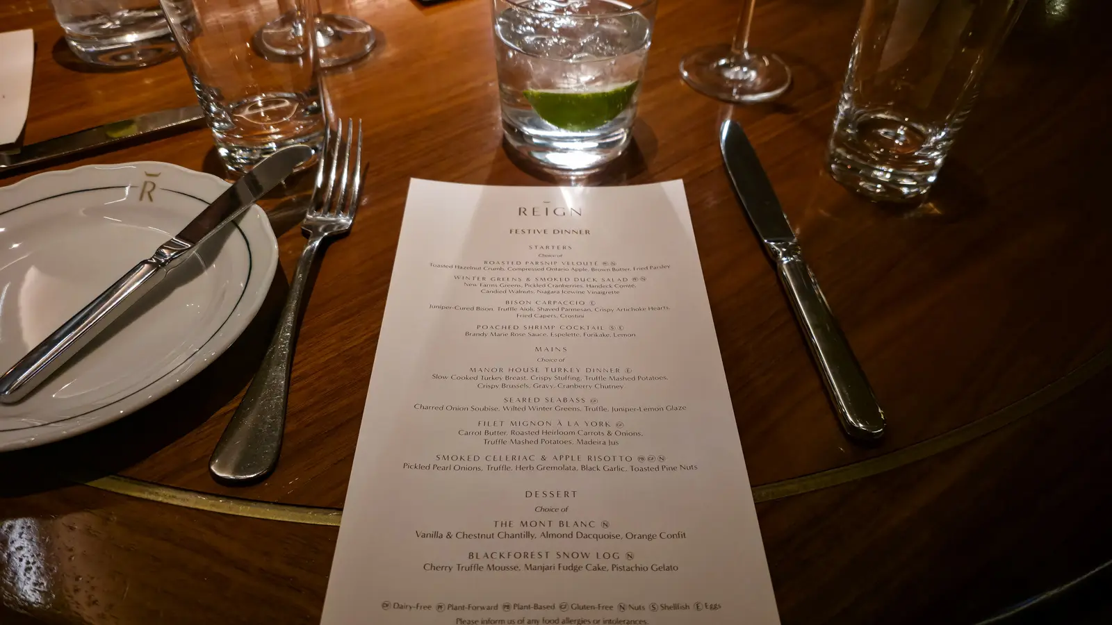 Reign Festive Dinner menu on a walnut table, with glasses and a plate marked with Reign's crowned R logo, Toronto