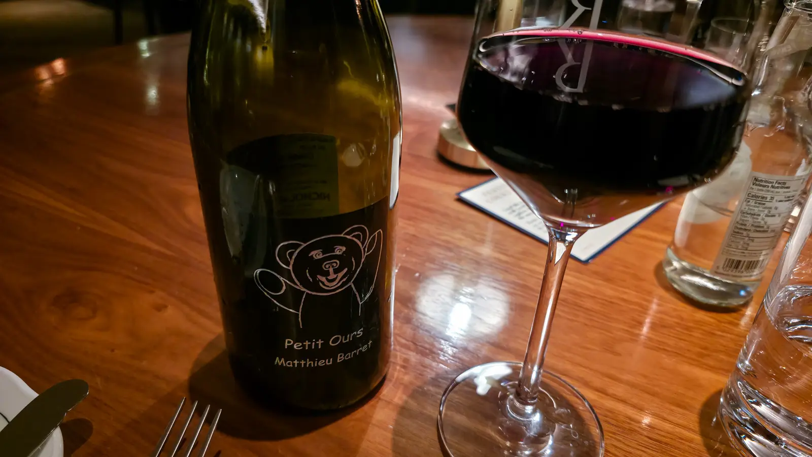 Bottle of Matthieu Barret Petit Ours with a bear drawing on the label, beside a full glass of red wine, Reign restaurant
