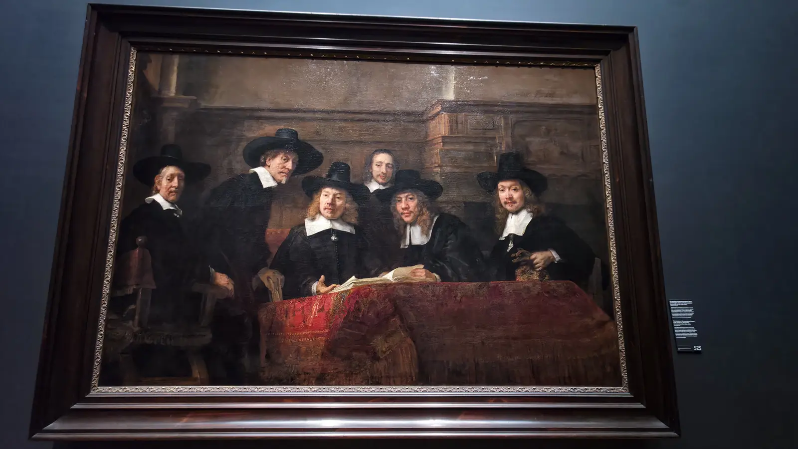 Rembrandt's The Syndics at the Rijksmuseum, five officials in black hats around a table with a red Persian rug and a servant behind