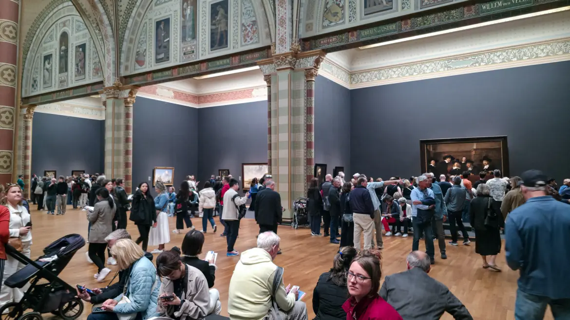 Crowds and benches in the Rijksmuseum's Gallery of Honour in Amsterdam, with Rembrandt's Syndics drawing a group on the right