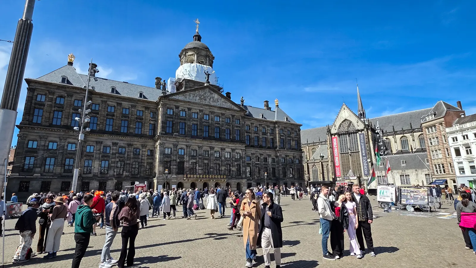 The Royal Palace and the Nieuwe Kerk on a crowded Dam Square, Amsterdam, under a clear blue sky