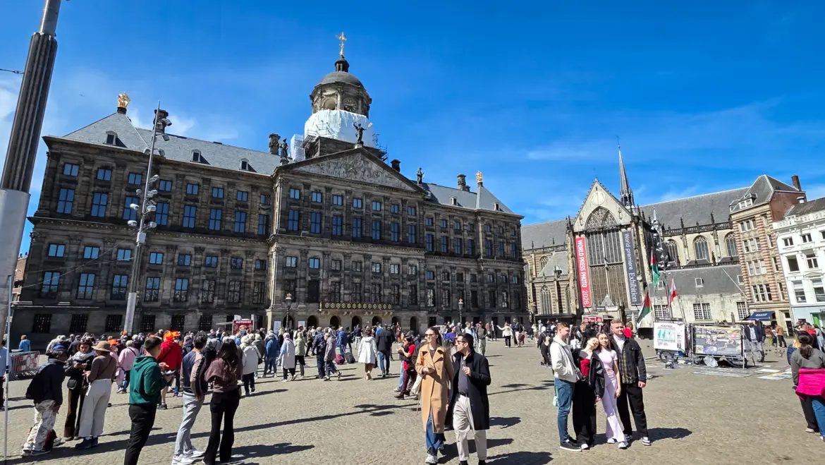 The Royal Palace and the Nieuwe Kerk on a crowded Dam Square, Amsterdam, under a clear blue sky