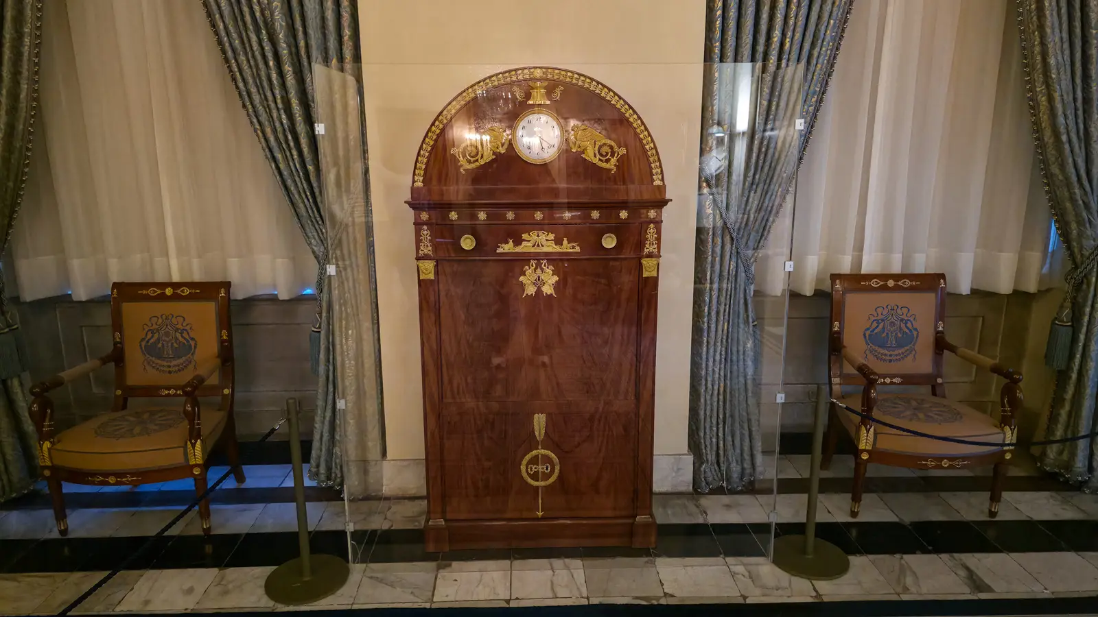 A tall mahogany secretaire with a clock in its arched top, gilt winged figures and an arrow keyhole, flanked by armchairs, Royal Palace Amsterdam