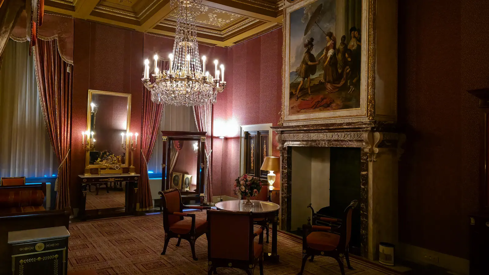 A deep red room in the Royal Palace Amsterdam with a chandelier, a gilt mirror and Theseus and Ariadne above the fireplace