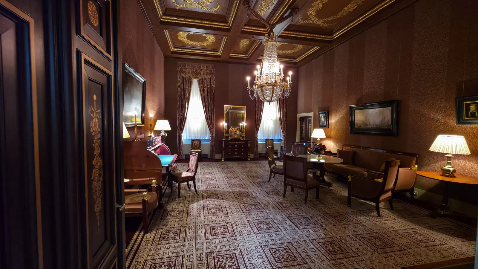 A long salon in the Royal Palace Amsterdam with a gilded coffered ceiling, crystal chandelier, patterned carpet and Empire furniture