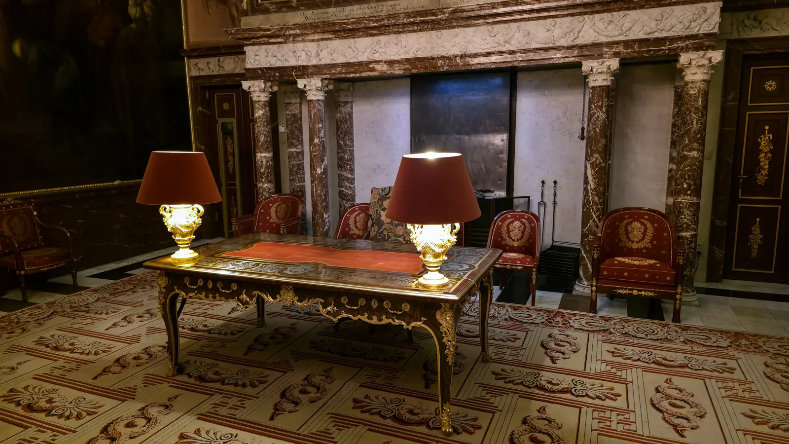 An inlaid desk with gilt mounts, a red leather top and two lamps in the Moses Hall of the Royal Palace Amsterdam, red armchairs behind