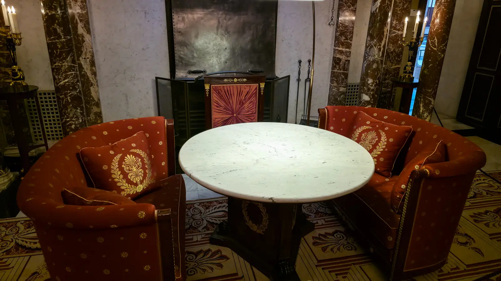 Two red Empire tub armchairs embroidered in gold around a white marble table in front of a fireplace in the Moses Hall
