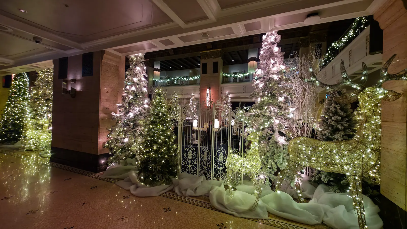 A pair of glittering wire reindeer and white ironwork gates among snowy, lit Christmas trees in the Fairmont Royal York lobby