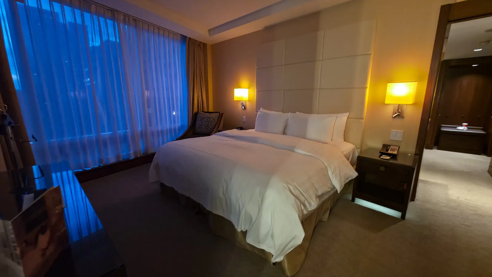 The king bed in a Shangri-La Toronto Deluxe Suite, with white linens, a tall padded headboard and wall lamps