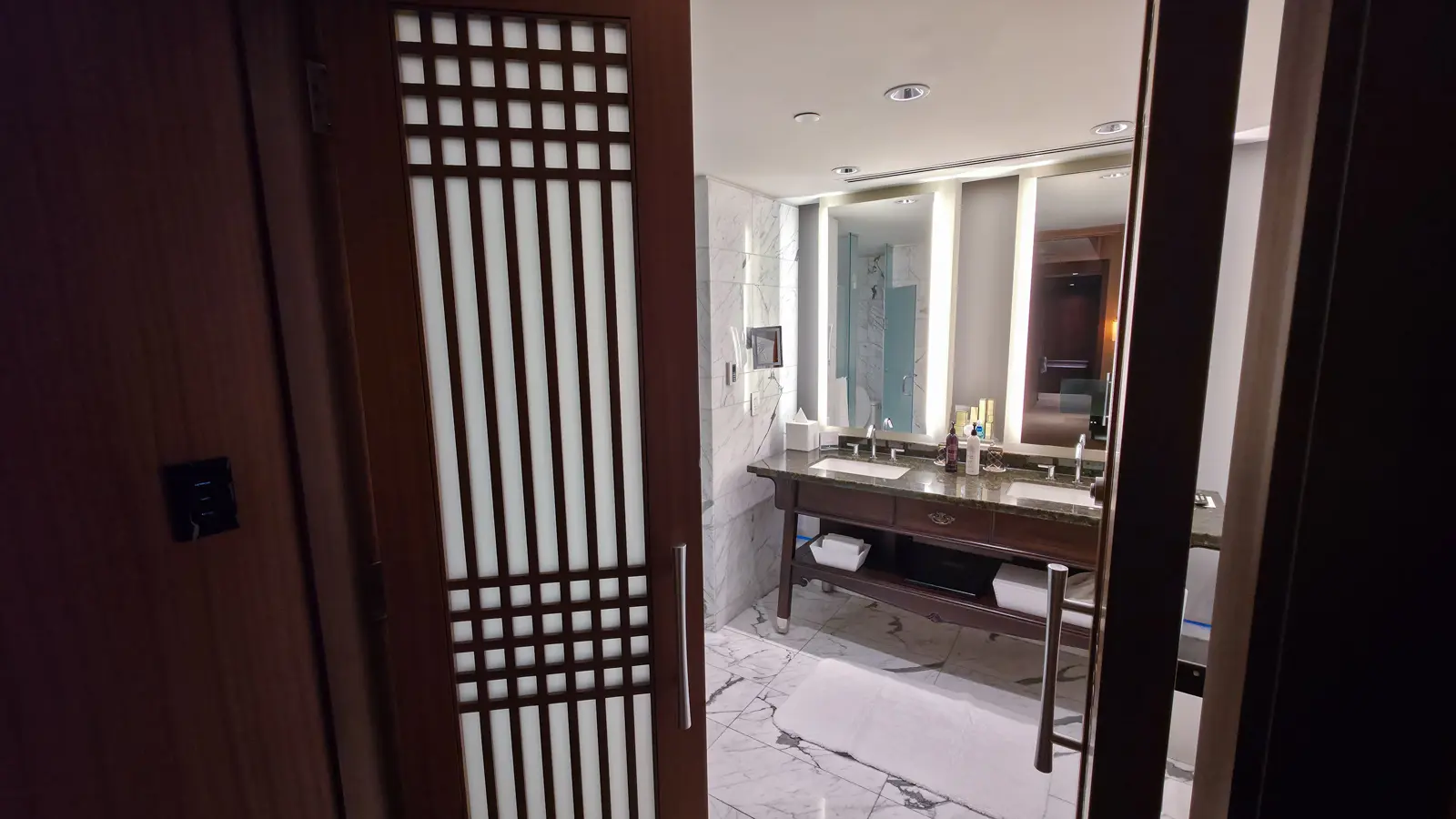 A wooden lattice door opening onto the marble master bathroom of a Shangri-La Toronto suite, with a double vanity and lit mirrors