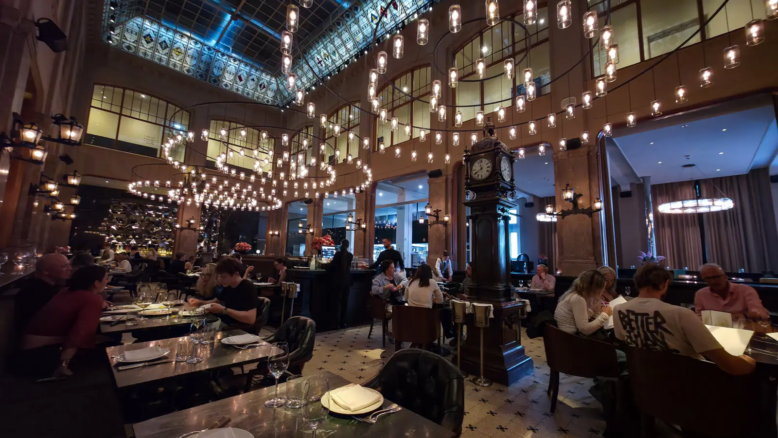 The Duchess dining room in Amsterdam, with rings of hanging bulbs under a glass roof and a standing clock among the tables