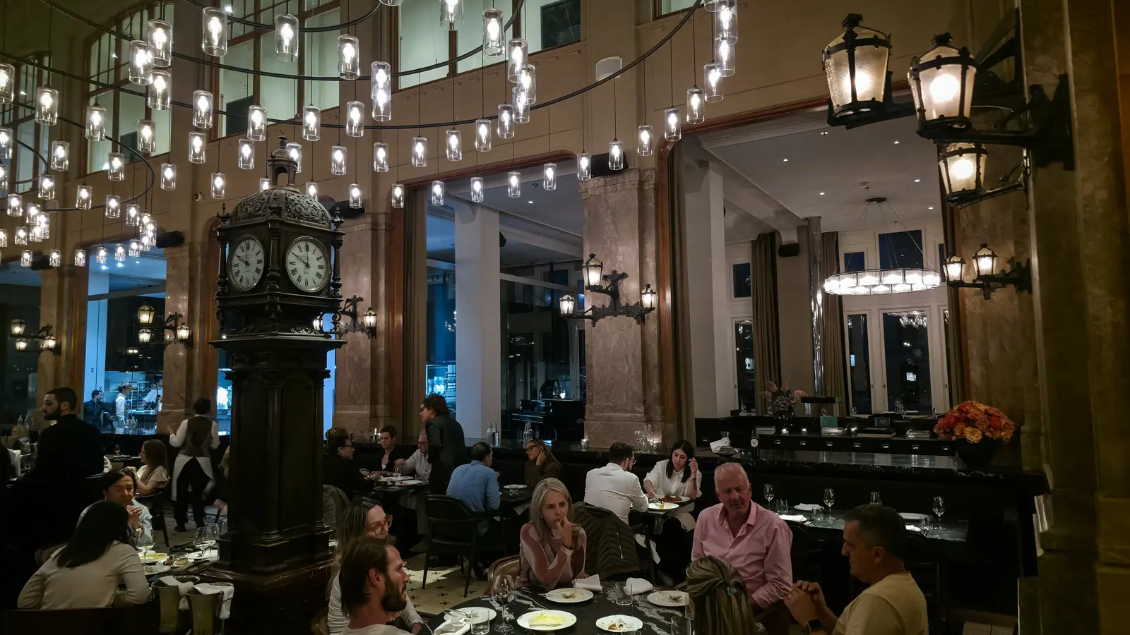 Diners around an ornate standing clock at The Duchess, with lanterns on the columns and the open kitchen behind glass