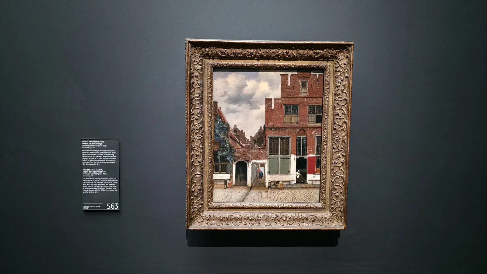 Vermeer's The Little Street in a carved gilt frame at the Rijksmuseum, two brick houses in Delft under a cloudy sky
