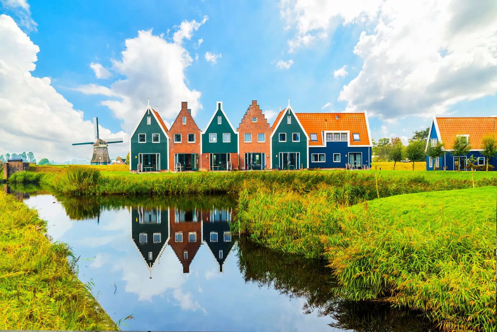 A row of green, red-brick and blue wooden houses in Volendam reflected in a pond, with a windmill on the left